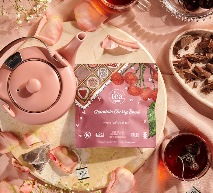 A pink teapot next to The Tea Spot's Chocolate Cherry Bomb tea package, surrounded by rose petals, a glass of tea, and broken chocolate pieces on a pink surface.