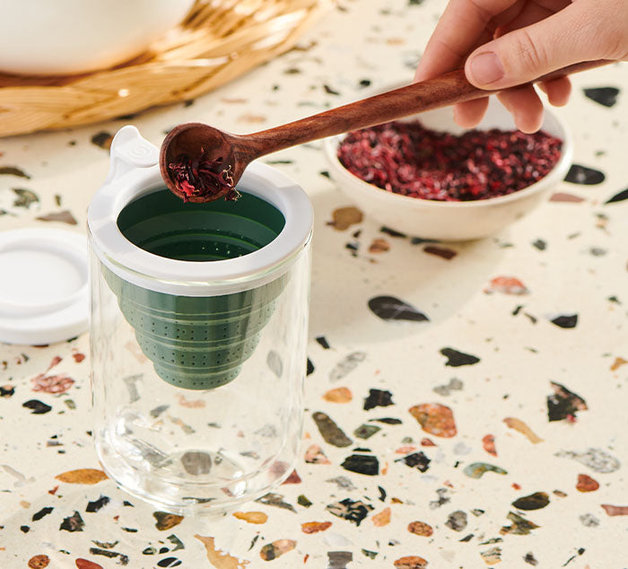 
                  
                    A hand spooning loose leaf tea into the green Tuffy Steeper, which is placed inside the clear double-walled Eclipse Mug, ready for brewing.
                  
                