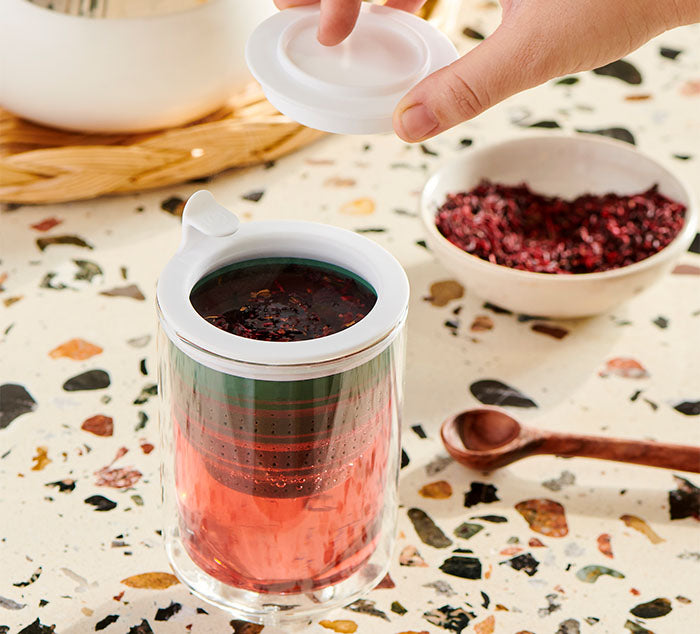 
                  
                    A hand placing the lid on the Eclipse Mug with a green Tuffy Steeper inside, as loose leaf tea steeps in hibiscus tea within the double-walled glass mug
                  
                