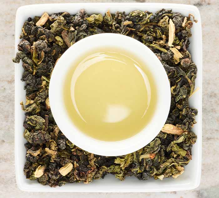 
                  
                    A small white cup of brewed Sugar Cookie Oolong tea centered on a square dish filled with loose tea leaves
                  
                