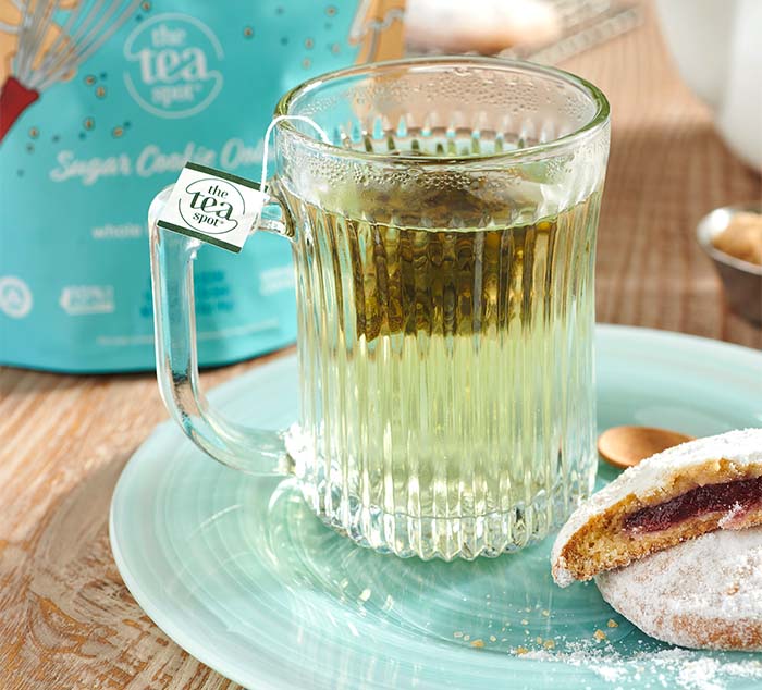 
                  
                    Glass mug of brewed Sugar Cookie Oolong tea on a blue plate, accompanied by a powdered cookie, with the tea packaging in the background
                  
                