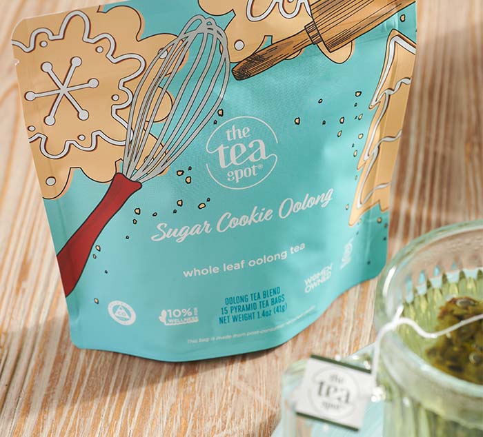 
                  
                    Packaging of The Tea Spot’s Sugar Cookie Oolong tea with festive baking illustrations, displayed on a wooden surface beside a glass mug of brewed tea
                  
                