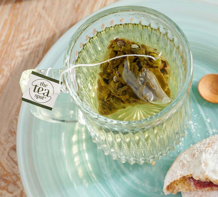 
                  
                    Glass mug with steeping Sugar Cookie Oolong tea sachet on a blue plate, accompanied by a powdered cookie and a small wooden spoon
                  
                