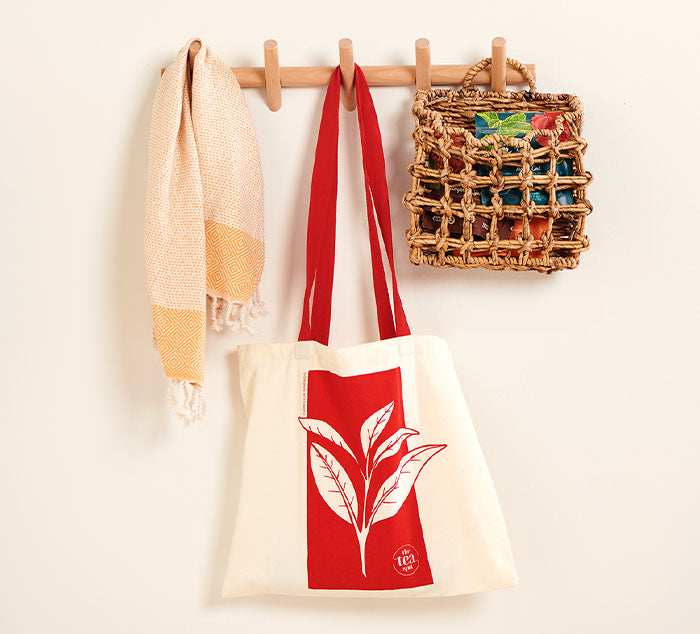 a white tote bag with a tea leaf design in red hangs on a wall next to a scarf and purse