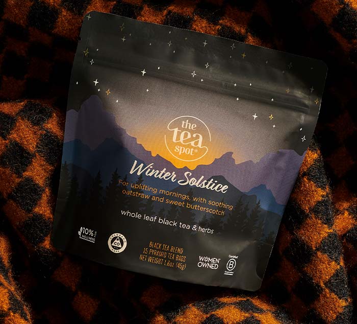 
                  
                    Winter Solstice Tea pouch resting on a warm, orange-and-black plaid blanket, showcasing its elegant packaging with mountain silhouettes, starry details, and a description of its soothing blend of oatstraw, butterscotch, and black tea
                  
                